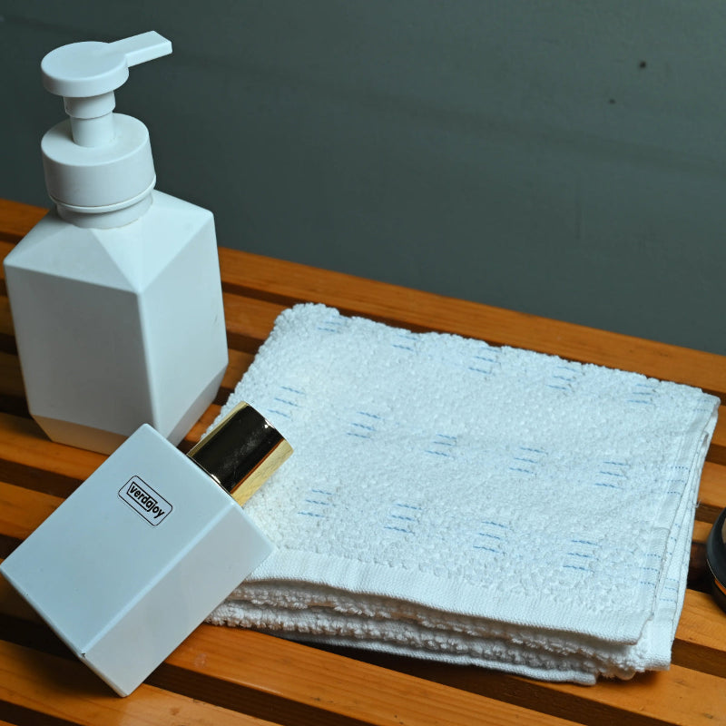 White soap dispenser, perfume bottle, and folded towel on a wooden surface with grey background.