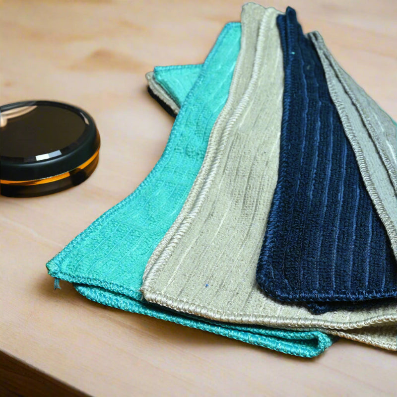 Set of folded cloths in teal, gray, and blue on a wooden surface with a plant in the background.